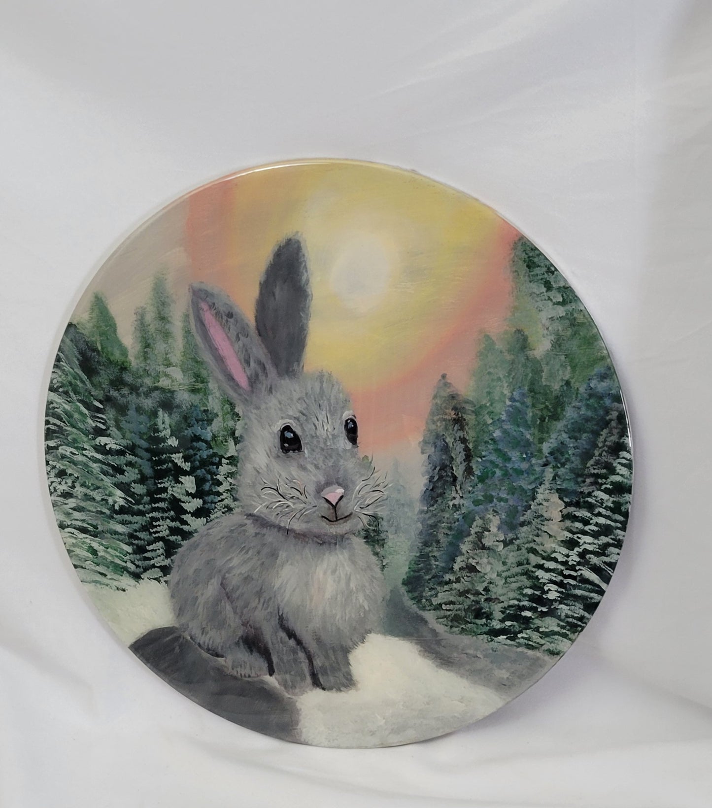 A hand painted birch wood round featuring a gray bunny with a snowy trail, frosted evergreens, and a sun-kissed twilight sky.