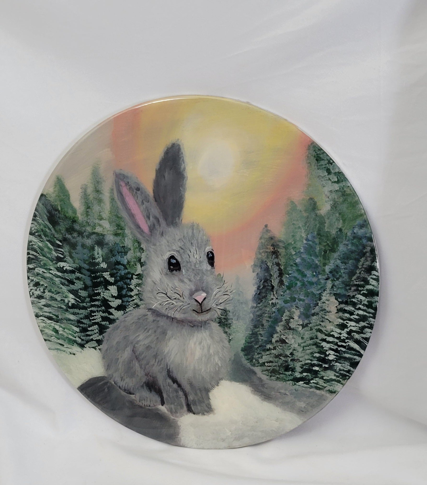 A hand painted birch wood round featuring a gray bunny with a snowy trail, frosted evergreens, and a sun-kissed twilight sky.